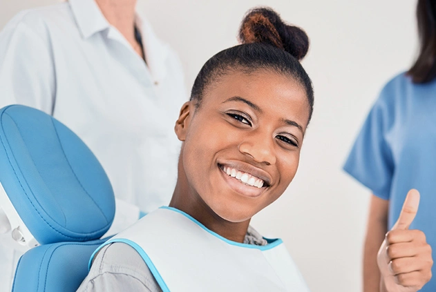 Smiling patient giving thumbs up in dental chair with dental professionals in background.