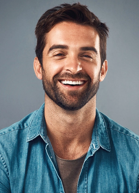 Smiling man in blue denim shirt against gray background, professional headshot for dental practice.