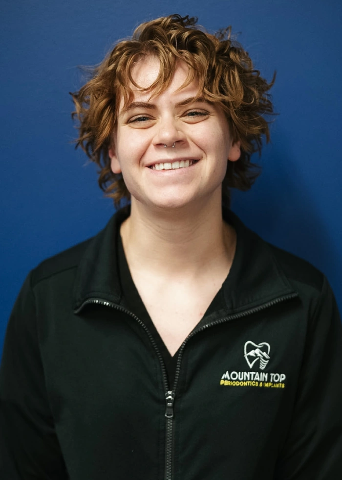 Professional headshot of smiling woman in black Mountain Top Periodontics jacket against blue background.