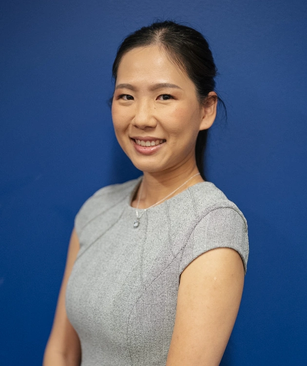 Professional portrait of smiling woman in gray top against blue background for dental practice website.