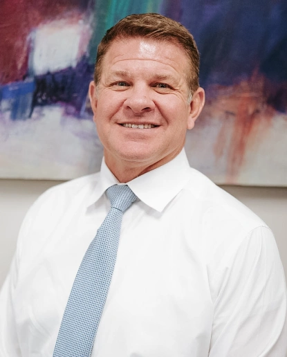 Professional headshot of male dentist in white shirt and blue tie against blurred office background.