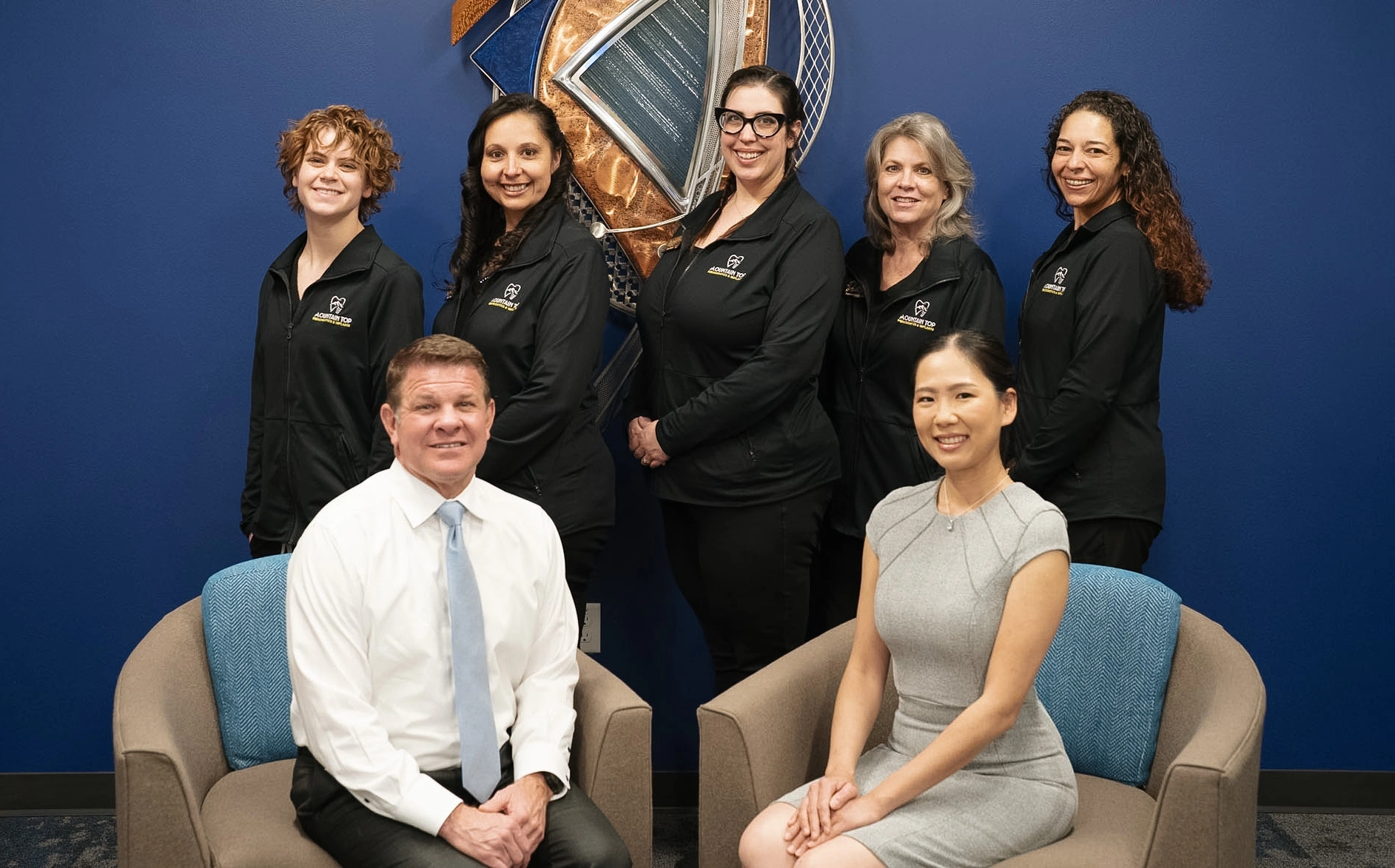 Professional dental team of eight people posing in office setting with blue backdrop and logo.