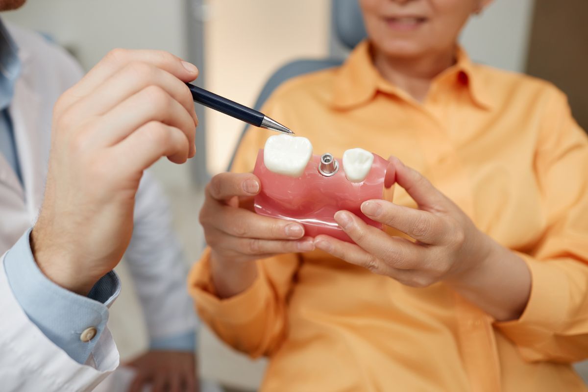 Dentist showing dental implant model with tooth replacement to patient in yellow shirt.